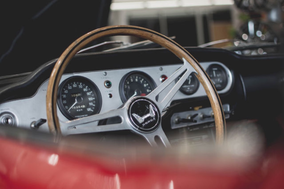 Wood-rimmed steering wheel and gauges of a vintage Honda, seen past a red fender